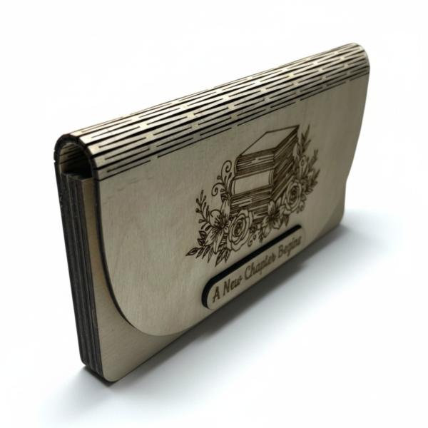 Holz-Umschlag aus hellem Birkensperrholz mit gelaserter Gravur A New Chapter Begins. Der Umschlag liegt dekorativ arrangiert, die natürliche Maserung und feinen Lasercut‑Details sind gut sichtbar. Ideal als Geschenkverpackung für Geld oder Gutscheine zu S